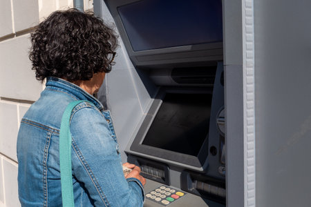 Woman is using an ATM machine. She is wearing a blue jacket. The ATM has a black screenの写真素材