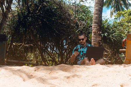 bearded man in sunglasses using laptop on sandy beach in tropicsの写真素材