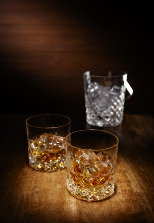A spotlight on a two crystal glass of scotch whisky with a ice bucket in the backghround, shot on a wooden table top.の写真素材