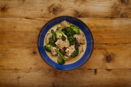 A blue bowl of succulent Thai Red Chicken Curry shot a wooden background with space for copyの写真素材