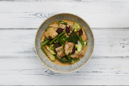 A bowl of succulent Thai Red Chicken Curry shot a white wooden background with space for copyの写真素材