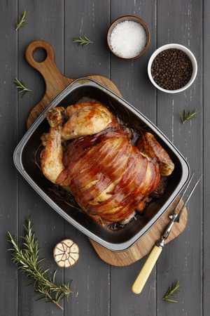 roast chicken in a rasting tin with ingredients and carving fork on a dark grey wooden backgroundの写真素材