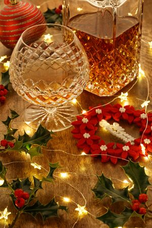 Crystal glass of brandy and a crystal brandy decanter with christmas lights and red christmas wreath decoration, on a wooden tableの写真素材