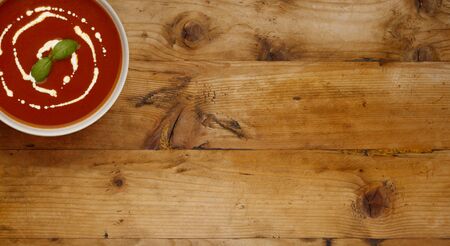 A  bowl of delicious tomato soup, topped with a drizzle of cream and a sprig of basil, in a rustic bowl, shot on a wooden background with space for copy.の写真素材