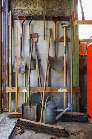 Old gardening tools including spades, brushes and watering can stacked against an old shed wall