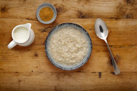 A nutritious bowl of porridge, a small bowl of brown sugar, a jug of milk and a silver spoon, on a checked wooden backgroundの写真素材