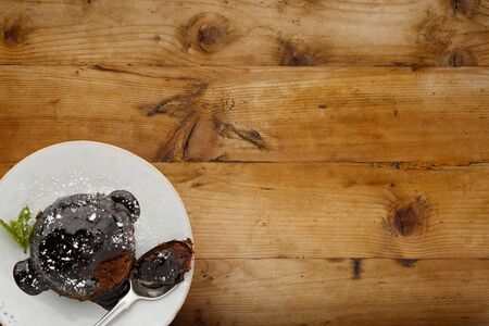 An individual chocolate sponge cake, with a sticky chocolate sauce, on a white plate with a spoon, shot on a wooden background with space for copyの写真素材