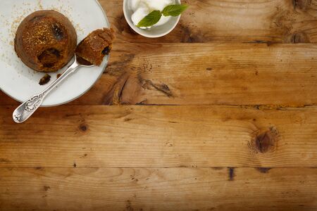 An individual spotted dick sponge cake, on a white plate with a spoon, and a small bowl of ice cream, on a wooden surface with space for copyの写真素材