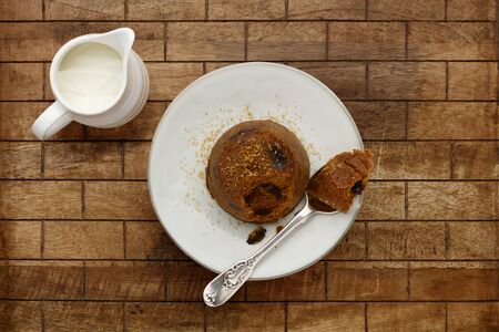 An individual spotted dick sponge cake, on a white plate with a spoon, and a small jug of cream, on a checked wooden surface with space for copyの写真素材