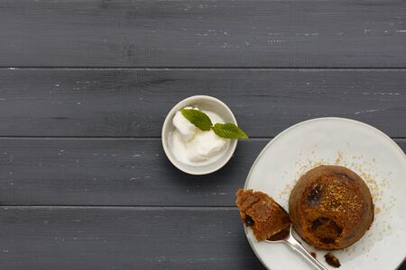 An individual spotted dick sponge cake, on a white plate with a spoon, and a small bowl of ice cream, on a dark grey wooden surface with space for copyの写真素材