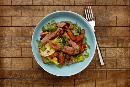 A delicious bowl of BBQ rump steak strips, sweet potatoe chips and vegetables, in a light blue bowl shot on a checked wooden background and forkの写真素材