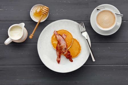 A plate of bacon and pancakes, drizzled with honey, a cup of coffee and jug of cream on a dark wooden backgroundの写真素材
