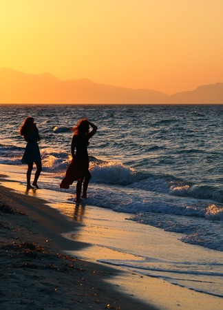 two young women in silhouette on a sunset beach one posing for a photo against a beautiful orange evening sky on the edge of breaking wavesの写真素材