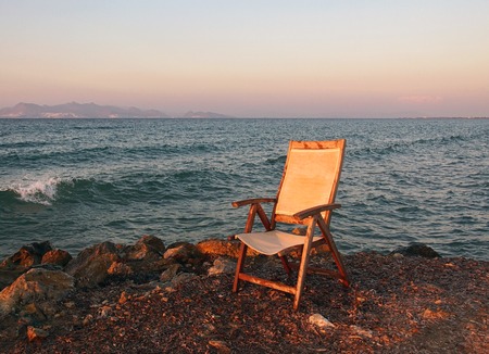 An old fading empty wooden chair in reflected sunset light left next to the sea on a calm summer eveningの写真素材