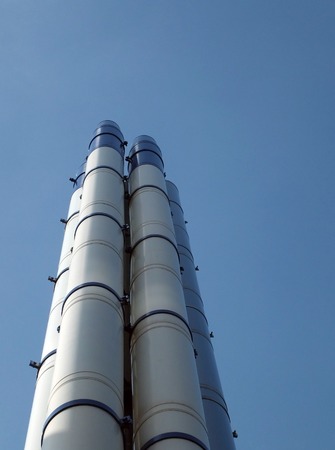 a group of tall modern white steel industrial chimneys standing against a bright blue sky and white...