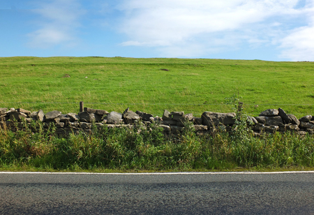 view of an empty country road with an overgrown dry stone wall with a fence separating it from a bright green hilly meadow with blue summer sky and white cloudsの写真素材