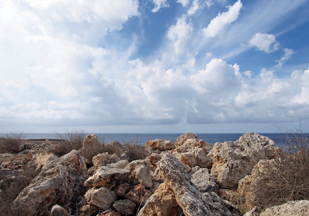 rough rocks and boulders with dry summer vegetation in front of a calm sea with white sunlit clouds filling the skyの写真素材
