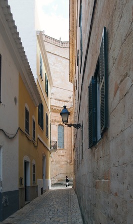 Cuitadella, Menorca, Spain - 3 October 2018: a woman walking past the end of a long quiet alley in cuitadella menorca with old historic buildings and the steps of the old cathedral in visible in the distanceの写真素材