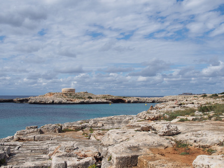 clifftop view of the coastline and the old british era Torre des Castellar at the edge of the bay in cala santandria near cuitadella with bright blue sea and summer cloudsの写真素材
