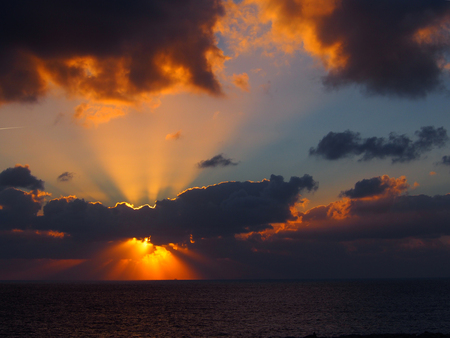 dramatic sunset over the sea with orange rays of light emerging from dark illuminated clouds over a calm oceanの写真素材