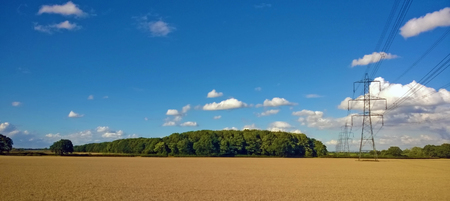 large electricity pylons and wires running though a flat sunlit wheat field with blue sunlit skyの写真素材