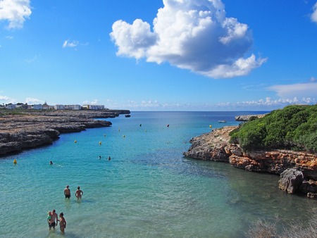 ciutaddal, menorca, spain - October 10 2018: view across the bay in cala santandria menorca with holidaymakers in the shallow sea with a bright blue sunlit skyのeditorial素材