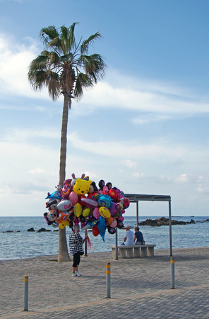 Paphos, Cyprus - March 17, 2018: a balloon seller walks past two tourists sat on the beach in Paphos cyprusのeditorial素材