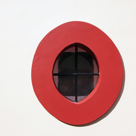 bright red stone oval window frame in a white wall with black metal bars in bright sunlightの写真素材