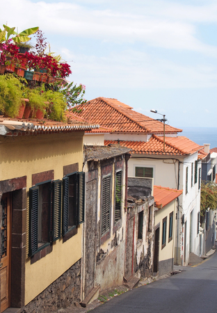 old traditional houses on a downhill street in funchal madeira with orange roof tiles and shuttered windows with colorful potted plants on the roof and the sea visible in the distanceの写真素材