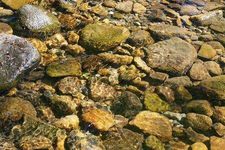 pebbles and rocks in a shallow stream with ripples reflecting sunlight in the waterの写真素材