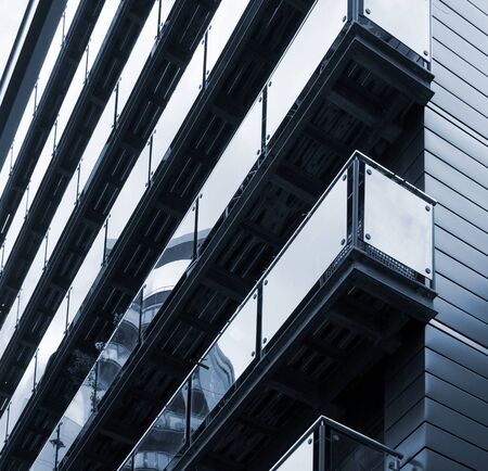 blue toned corner view of modern apartments with stylish glass paneled balconies reflect nearby urban buildingsの写真素材