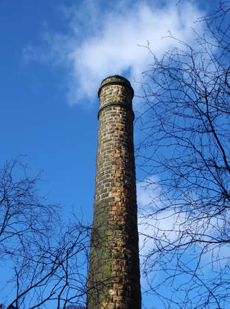 vertical view of an old stone chimney in a ruined mill surrounded by trees against a blue cloudy sky in the Colden Valley west yorkshireの写真素材