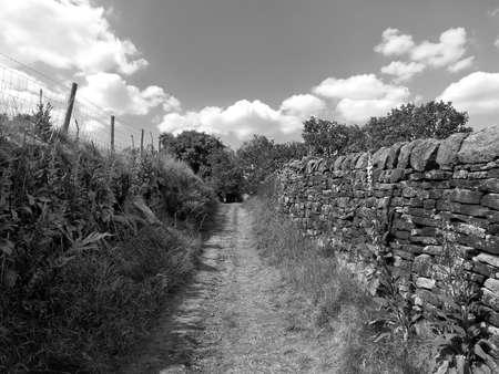monochrome perspective view of a long narrow country lane running uphill surrounded by high stone...