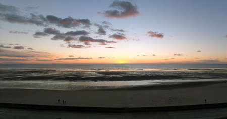 wide panoramic twilight beach scene with darkening pink sunset sky and clouds reflected in a calm sea people and water on the beachの写真素材