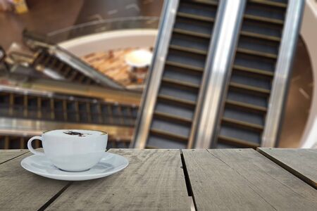 Defocus with terrace wood and cappuccino coffee with escalators in shopping mall for background...