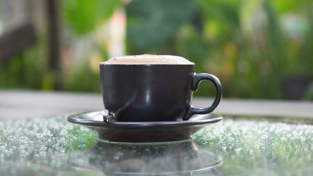 Hot cappuccino coffee cup on grass table in garden with dewdrop foregroundの写真素材