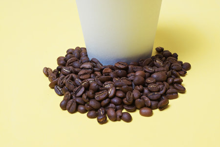 coffee beans next to a paper cup on a white-yellow backgroundの写真素材