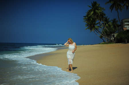 A woman of 30-40 years old in a white lace dress stands on the ocean, and waves of clear water reach her feet with a sea of different colors from turquoise to darkの写真素材