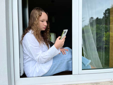 a girl in a white shirt a teenager sits on a white windowsill the view from the street she can be...