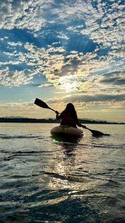 teenage girl is kayaking at sunset in Pacific Ocean, only the silhouette of Kayak Paddles is visible she swims along sunny path to the sea. a beautiful sky with small clouds and the sun shines throughの写真素材