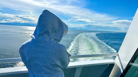 teenage girl sails away on liner she looks into distance she is dressed in a gray hoodie with her back to camera against the background of the blue sky and the blue ocean Ferry footprints on the seaの写真素材