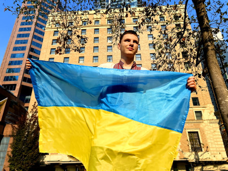 A Ukrainian guy in an embroidered shirt pounces on the shoulders of Ukrainian flag he stands against the backdrop of building and asks for help Fight against Russian terrorism Ukrainians in Canadaの写真素材