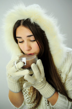 New Year and Christmas Party Invitation Delicate beauty joy of life Beautiful young girl drinks from a glass cup of coffee or cocoa White coat and white gloves Snow Maiden with long hair put on a hoodの写真素材