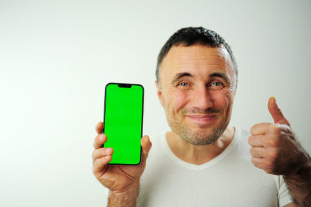 Positive millennial man showing huge brand new smartphone with empty screen and thumb up over white studio background, mockup, panorama with copy space, newest mobile app conceptの写真素材