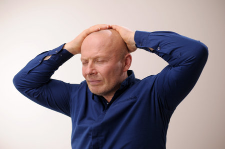 Need medicine for migraine Headache man put his hands on his head and closed his eyes an unpleasant sensation Emotions of an adult handsome bald man A man on a white background in a blue shirtの写真素材