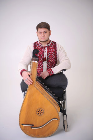 handsome young man with kobza musical instrument in hands sits on white background on wheelchair without legs war veteran dressed in Ukrainian embroidered shirt national symbols of Ukraine vyshyvankaの写真素材