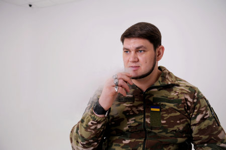 Adult handsome man 35 years old Military man in uniform sitting and smoking He exhales puffs of smoke He has rings on his fingers emblem of Ukraine The Ukrainian flag is sewn on the protective jacketの写真素材