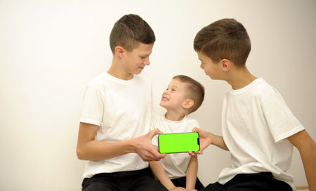 Three brothers with smartphone three boys in white t-shirts sit hold a phone with a green chromakey screen in front of them. They look at each other joyfully. advertising space for text. teenagerの写真素材
