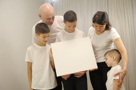5 people three boys mom and dad Beautiful smiling family in white T-shirts hold in hands the big blank banner and Look at the paper posterの写真素材