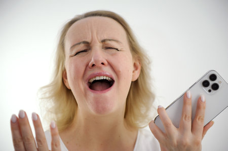 white background close-up woman screaming with mouth open holding the phone in hands raised hands and spread her fingers to the sides joy dissatisfaction success victory defeat good news bad newsの写真素材